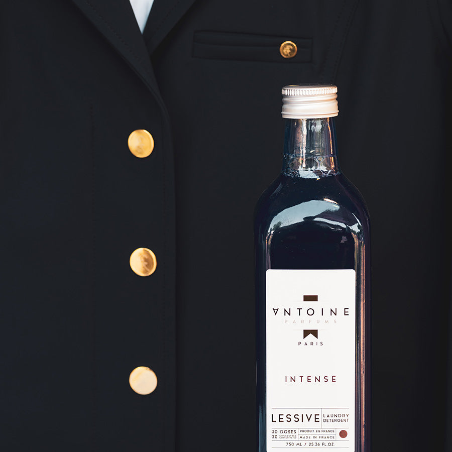 Antoine Intense perfumed laundry detergent in a luxurious glass bottle, displayed against a black suit backdrop.