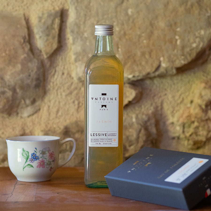 Antoine Jasmin perfumed laundry detergent in a glass bottle, displayed on a wooden table with elegant dishware, evoking a fresh, floral ambiance.