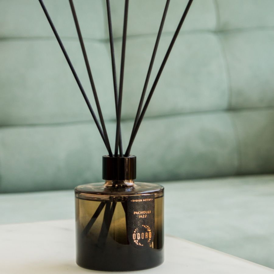A home fragrance bottle elegantly displayed on a table, with a blurred green sofa in the background, adding a touch of color and sophistication to the cozy interior setting.