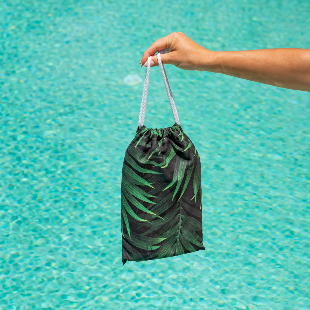 Hand holding Jungle Drift towel pouch over turquoise pool water – lightweight and easy to carry.