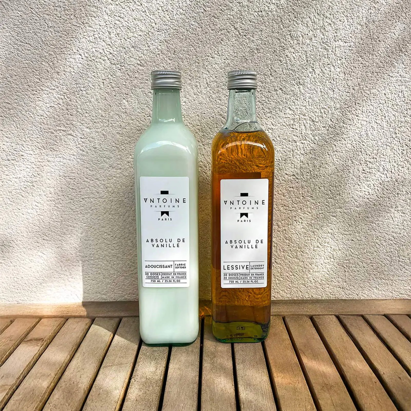 Antoine Parfums Absolu de Vanille laundry detergent and fabric softener bottles standing on a wooden table in natural sunlight – French luxury home fragrance set.
