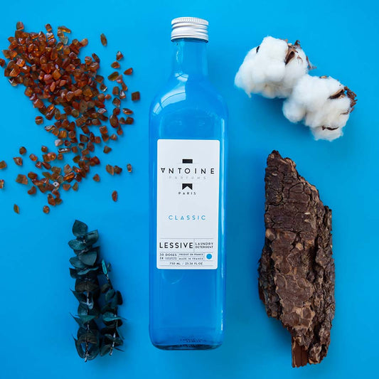 Antoine Classic perfumed laundry detergent in a glass bottle, surrounded by natural ingredients on a blue background.